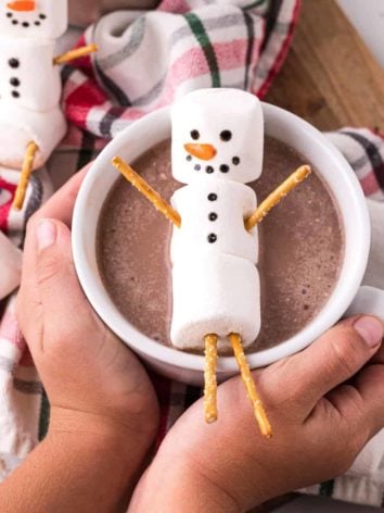 Marshmallow snowmen, one atop a cup of cocoa cupped between two hands.