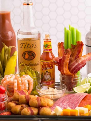 Assorted ingredients to make bloody marys on a board including bacon and vodka.