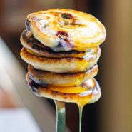 Stack of small blueberry pancakes on a fork, dripping syrup.